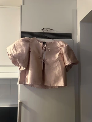 Ljusrosa blus med puffärm från Gina Tricot - Supersöt ljusrosa blus från Gina Tricot med glansigt satinmaterial och stora puffärmar. Blusen har rund halsringning och knytband framtill som ger en extra gullig touch. Perfekt för dig som gillar statement-ärmar och en feminin vibe. Aldrig använd!