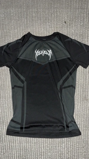 Svart t-shirt YOUNGLA - Svart teknisk t-shirt från YOUNGLA med gråa grafiska detaljer och logga i vitt framtill. T-shirten har korta ärmar och är gjord i ett lätt och ventilerande polyestermaterial som passar perfekt för träning. Snygg, sportig och modern design. Använd nån få gång