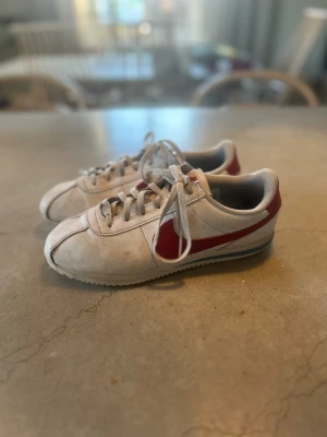 Nike Cortez vita och röda sneakers - Klassiska Nike Cortez sneakers i vitt med röd swoosh och häl, samt blå detalj på sulan. Skorna har snörning och en retro vibe med låg profil och rund tå. Perfekta för dig som gillar old school streetstyle och vill sticka ut med en ikonisk modell.