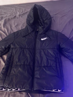 Svart pufferjacka från Nike med huva - Svart pufferjacka från Nike med vit logga på bröstet, huva med snörning och dragkedja framtill. Jackan har en clean look och är perfekt för kalla dagar. Materialet är syntet och jackan har en loose passform som ger en chill vibe.