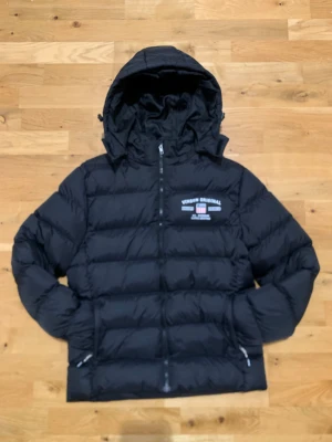 Svart pufferjacka från Vinson Original - Varm och skön Svart pufferjacka från Vinson Original med avtagbar luva och två sid fickor med dragkedjor, jackan har även en ficka inuti. Den är i väldigt bra skick och är nästan som ny. Storleken är 160 