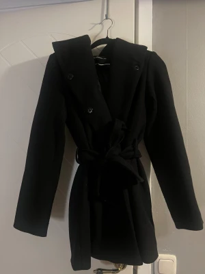 Svart kappa med huva och bälte - Snygg svart kappa med stor huva och bälte i midjan. Dubbelknäppt framtill med svarta knappar och långa ärmar. 