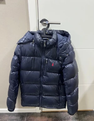 Mörkblå pufferjacka från Polo Ralph Lauren - Snygg mörkblå pufferjacka från Polo Ralph Lauren med huva och dragkedja framtill. Jackan har en röd Polo-logga på bröstet, flera praktiska fickor med dragkedja och en quiltad design som ger en cool look. Perfekt för kalla dagar och enkel att matcha med din stil. Priset går att diskutera 