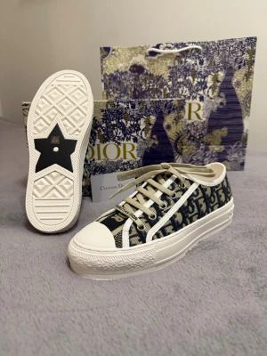 Dior sneakers med monogram och snörning - Snygga Dior sneakers med beige och marinblått monogrammönster, platt vit sula och rund tå. Skorna har beige snörning och detaljer i textil, med tydlig Dior-logga på sidan och sula. Perfekt för dig som vill sticka ut med lyxig streetstyle.