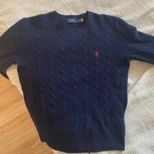 Marinblå kabelstickad tröja Polo Ralph Lauren - Snygg marinblå kabelstickad tröja från Polo Ralph Lauren med klassisk röd broderad logga på bröstet. Rund halsringning och ribbade muddar. Perfekt för dig som gillar stilrena och tidlösa plagg med premiumkänsla.