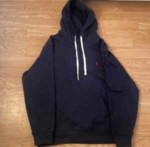 Mörkblå hoodie från Polo Ralph Lauren - Snygg mörkblå hoodie från Polo Ralph Lauren med vit snörning och klassisk röd logga broderad på bröstet. Tröjan har huva, känguruficka och ribbade muddar. Perfekt för chill dagar och enkel att matcha med jeans eller joggers.