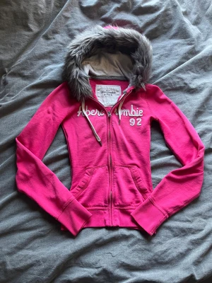 Rosa hoodie med päls från Abercrombie - Snygg rosa hoodie från Abercrombie & Fitch med grå fuskpäls runt huvan och broderad logga på bröstet. Tröjan har dragkedja, snörning vid huvan och två fickor framtill. Perfekt för dig som gillar streetstyle och vill ha något varmt och chill.omsydd till xxs