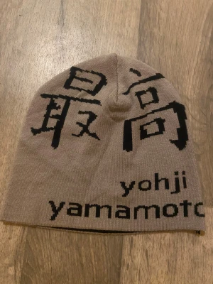 Supreme x Yohji Yamamoto mössa - Säljer en Supreme x Yohji Yamamoto mössa med svart grafiskt tryck och japanska tecken. Mössan är stickad i mjukt material och har Supreme-loggan på ena sidan och Yohji Yamamoto på den andra. Perfekt för dig som gillar streetwear och unika designer. 