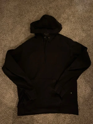 Svart hoodie från Hugo Boss, strl M - Svart hoodie från Hugo Boss med broderad logga på bröstet. Tröjan har huva med snörning, ribbade muddar och en stor magficka. Tillverkad i mjuk bomull och polyesterblandning, perfekt för chill dagar. Snygg och stilren design som passar till streetwear.