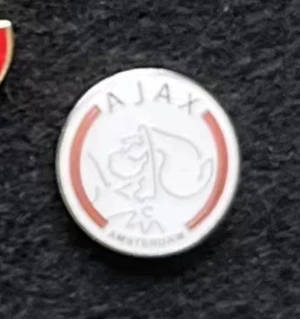Ajax Amsterdam pin vitt och rött - Snygg rund brosch med Ajax Amsterdam-loggan i vitt och rött. Den  har en stilren design med klubbens namn och en grafisk profil av Ajax-hjälten. Perfekt för dig som älskar fotboll och vill visa ditt stöd för laget. Den är i metall Passar att sätta på jacka, väska, kavaj eller keps!  