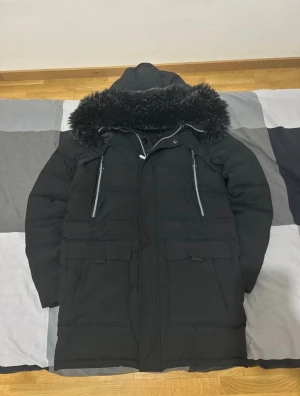Svart lång parkas med fuskpäls - Säljer en svart lång parkas från supply demand med stor huva och svart fuskpäls runt kanten. Jackan har dragkedja och knappar framtill, två stora fickor med lock och justerbara snören vid huvan. Perfekt för kalla dagar och riktigt snygg streetstil. 