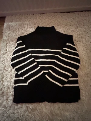 Randig svartvit polotröja Gina Tricot - Svart stickad polotröja från Gina Tricot med vita horisontella ränder. Tröjan har hög krage och långa ärmar, perfekt för kyliga dagar. Klassisk och stilren design som funkar till många outfits.