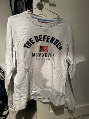 Vit sweatshirt från The Defender - Vit sweatshirt från The Defender med broderad text och flagga på bröstet. Rund halsringning och långa ärmar. Premium Collection.