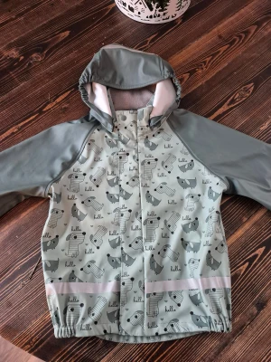 Grå regnjacka med hundmönster Ocean Works - Grå regnjacka från Ocean Works i storlek 122-128. Jackan har ett gulligt hundmönster, avtagbar huva och reflexdetalj nedtill. Tillverkad i vattenavvisande material. Perfekt för blöta dagar och håller dig torr med stil.
