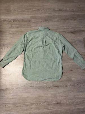 Grön skjorta från Polo Ralph Lauren - Snygg ljusgrön skjorta från Polo Ralph Lauren med klassisk button-down krage och broderad logga på bröstet. Skjortan har långa ärmar, knäppning framtill och är tillverkad i mjuk bomull. Perfekt för dig som gillar stilrena och tidlösa plagg. En knapp är borta på toppen på vänster sida.