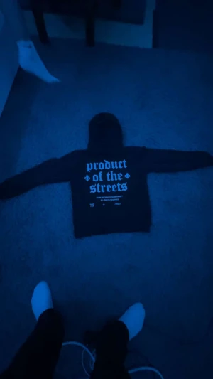 Svart hoodie från Hoodrich med tryck - Svart hoodie från Hoodrich med stor vit text 'HOODRICH' på bröstet och broderade detaljer i rött och blått. På ryggen står det 'product of the streets' i vit text. Tröjan har huva och lång ärm, tillverkad i mjuk bomull för skön komfort.