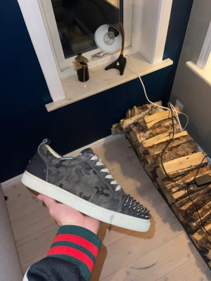 Christian Louboutin sneakers - Säljer ett par svarta och grå sneakers från Christian Louboutin med coolt kamouflagemönster i mocka och skinn. Tåpartiet är täckt av silvriga nitar för en edgy vibe. Skorna har vita snören och vit platt sula. Perfekta för dig som vill sticka ut. Dustbags fås med 