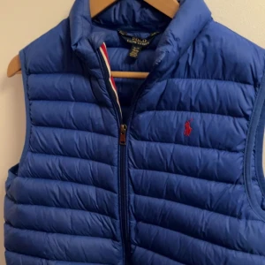 Blå väst från Polo Ralph Lauren - Säljer en snygg blå dunväst från Polo Ralph Lauren med dragkedja och röd logga på bröstet. Perfekt för kyligare dagar och enkel att matcha med olika outfits. Funkar även under vinter jacka eller dunjacka XL 18-20 motsvarar mellan XS - S. Sällsynt, 8/10 skick då det finns små tecken av användning om man kollar noggrant nära men inga defekter , pris går alltid att diskutera! ✅