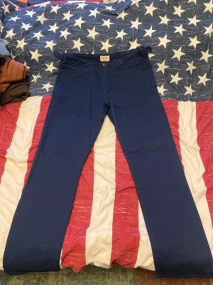 Mörkblå chinos från East West - Snygga mörkblå chinos från East West med raka ben och klassisk passform. Byxorna har två fickor fram och två bakfickor med lock och knapp, samt justerbara knappar i midjan. Perfekta för en clean och stilren look. Byxorna är knappt använda men de är inte helt nya. Storlek 33/32. 