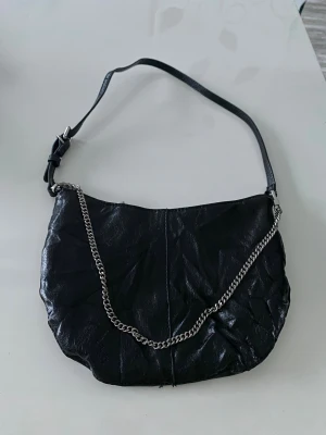 Svart axelväska med kedja från Zara - Snygg svart axelväska från Zara i skinnimitation med cool silverfärgad kedjedetalj framtill. Väskan har justerbar axelrem och dragkedja upptill. Insidan är fodrad i grått tyg och har en innerficka med dragkedja. Perfekt storlek för det viktigaste.
