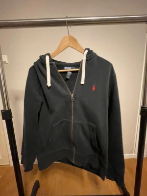 Svart hoodie från Polo Ralph Lauren - Svart hoodie med dragkedja från Polo Ralph Lauren. Tröjan har vit snörning i huvan, klassisk röd broderad logga på bröstet och en stor ficka framtill. Materialet är 84% bomull och 16% polyester, vilket gör den mjuk och skön. Perfekt för chill dagar.