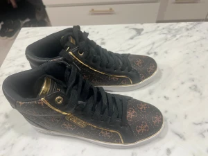 Svarta sneakers från Guess med guld - Snygga svarta sneakers från Guess med brunt monogrammönster och gulddetaljer. Skorna har svarta snören, rund tå och vita sulor. Materialet är syntet med läderlook och inslag av guld  känsla. Helt nya  aldrig använda  Säljes för 500kr 