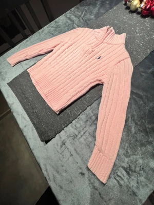 Rosa stickad polotröja från Ralph Lauren - Supersnygg rosa stickad polotröja från Ralph Lauren med klassisk kabelstickning och broderad logga på bröstet. Tröjan har lång ärm, ribbade muddar och en hög krage som ger en clean och preppy vibe.
