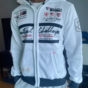 Vit hoodie Geographical Norway - Vit hoodiejacka från Geographical Norway med broderade flaggor, stora tryck och detaljer i svart och rött. Jackan har dragkedja, huva och ribbade muddar. Snygga broderier och tryck både fram och bak, samt fickor framtill. Storlek M