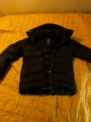Svart dunjacka från Haglöfs - Svart dunjacka från Haglöfs med huva och diskret logga på ärmen. Jackan har quiltad design, dragkedja och knappar framtill. Perfekt för kalla dagar med sin fluffiga och stilrena look. Passar dig som vill ha en varm och snygg jacka i vinter. Orginal pris: 2500-3000kr. Pris kan diskuteras 