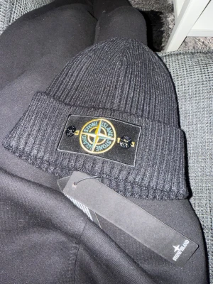 Svart ribbad mössa från Stone Island - Svart ribbstickad mössa från Stone Island med klassisk logotyp framtill, fäst med två knappar. Tillverkad i 100% bomull och har en snygg uppvikt kant. Perfekt för dig som vill ha en clean och stilren look med street-vibe.