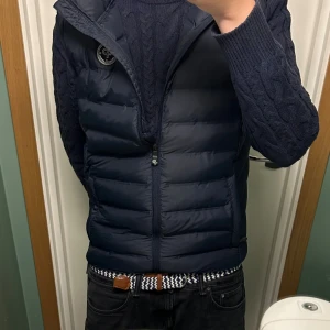 Mörkblå dunväst från Pelle P - Snygg mörkblå dunväst från Pelle P med quiltad design och dragkedja framtill. Västen har ett rundat emblem på bröstet och praktiska sidofickor med dragkedja. Perfekt att slänga över en hoodie eller stickad tröja för extra värme.