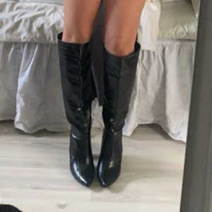 Svarta höga stövlar med klack - Säljer ett par svarta stövlar med spetsig tå och hög klack. Stövlarna är tillverkade i glansigt skinnmaterial och går upp till knäna. Snygg och stilren design som ger en edgy vibe till din outfit.