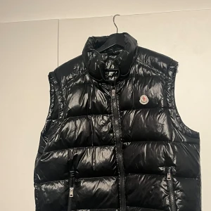 Svart dunväst från Moncler - Svart, glansig dunväst från Moncler med hög krage och två dragkedjefickor framtill. Västen har quiltad design och klassisk Moncler-logga på bröstet. Perfekt för lager-på-lager och har en cool, urban vibe. Det går att skanna både märket där fram och koden i västen! 