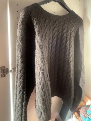 Svart stickad tröja med kabelmönster - Mysig svart stickad tröja med klassiskt kabelmönster. Tröjan har rund hals och långa ärmar, perfekt för kyliga dagar. Materialet känns mjukt och värmande, och den har en loose passform som gör den extra skön att bära.