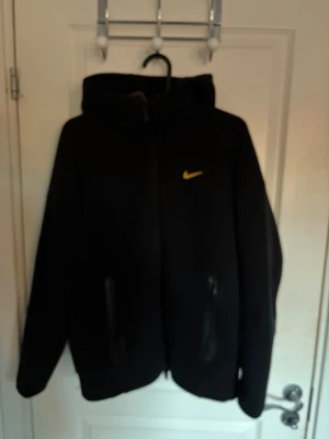Svart Nike vindjacka med huva Nocta - Säljer en svart vindjacka från Nike med gul logga på bröstet. Jackan har huva, hel dragkedja framtill och två fickor med dragkedja. Perfekt för dig som vill ha en enkel och stilren jacka med sportig vibe.