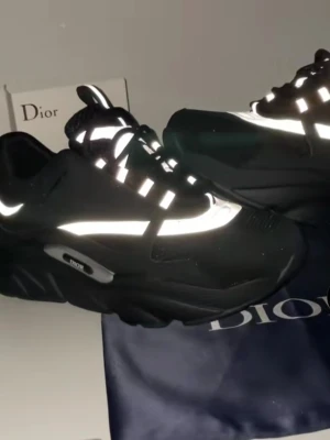 Svarta Dior sneakers med reflekterande detaljer - Säljer ett par svarta Dior sneakers med chunky sula och reflekterande vita detaljer. Skorna har snörning, meshpaneler och logotyp på sidan. Snygg och modern design som sticker ut med de reflekterande inslagen. Perfekt för dig som gillar exklusiva och trendiga sneakers.