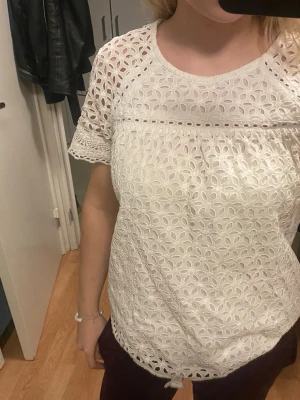 Vit blus med spetsdetaljer - Säljer en vit blus med korta ärmar och rund hals. Blusen har ett snyggt broderat hålmönster över hela tyget och spetsdetaljer vid ärmslut och nederkant. Perfekt för dig som gillar ljusa och luftiga toppar med fina detaljer.