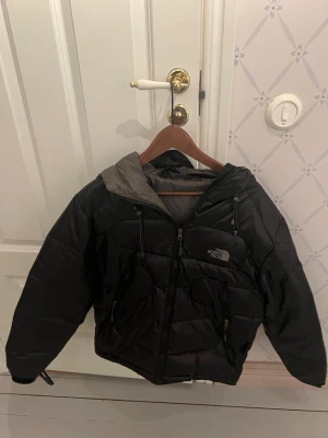 Svart pufferjacka från The North Face - Svart pufferjacka från The North Face med huva och dragkedja framtill. Jackan har två stora fickor med dragkedja och justerbara snören vid huvan. Insidan är grå och materialet är glansigt syntet. Perfekt för kalla dagar och har en riktigt chill vibe.             