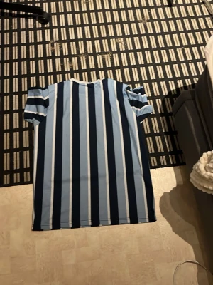 Randig t-shirt från Ellesse - Säljer en fräsch t-shirt från Ellesse med vertikala ränder i ljusblått, marinblått och vitt. T-shirten har korta ärmar, rund hals och ett broderat Ellesse-logo på bröstet. Materialet är mjuk bomull som känns skön mot huden. Perfekt för dig som gillar sportig och stilren look.