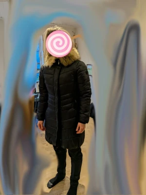 Svart lång dunjacka med fuskpäls - Säljer en svart lång dunjacka från Didriksons med huva och stor fuskpäls runt kanten. Jackan är quiltad, har dragkedja framtill och två fickor med dragkedja. Perfekt för kalla dagar och riktigt mysig att bära.