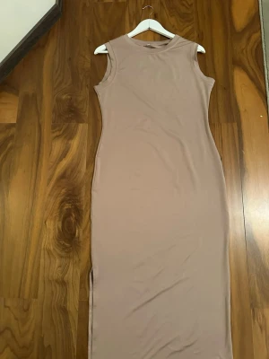 Beige långklänning med slits - Säljer en stilren beige långklänning i mjukt material. Klänningen är ärmlös och har en rund halsringning. Den är figurnära och har en snygg slits på sidan för extra rörelsefrihet. Perfekt för dig som gillar minimalistisk och enkel design.