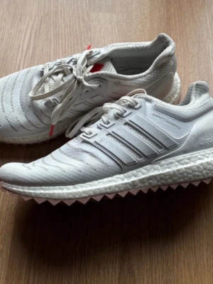 Adidas Ultraboost vita sneakers strl 40,5 - Säljer ett par Adidas Ultraboost GX6848 sneakers i vitt med klassiska tre ränder på sidan och snörning. Ovandelen är i mesh och sidorna har detaljer i syntet. Sulan är boost och yttersulan har ett mönstrat grepp i ljusbrunt. Perfekta för träning eller chill.