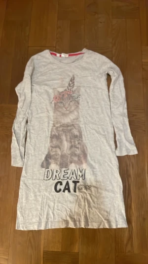 Ljus långärmad kattklänning bpc - Långärmad ljus natt klänning från bpc bonprix collection med ett stort kattmotiv och texten 'DREAM CAT' framtill. Klänningen har rund halsringning och är gjord i mjuk bomull. Perfekt för dig som gillar gulliga prints och avslappnad stil. Storlek 152/158. Pyjamas. Pris kan diskuteras. Väl använd.