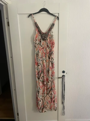 Mönstrad långklänning med pärldetaljer - Säljer en bohemisk långklänning med färgglatt mönster i vitt, rosa, beige och brunt. Klänningen har djup v-ringning med pärldekorationer och är ärmlös med smala axelband. Perfekt för sommarens varma dagar och har en flowy passform.