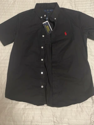 Svart kortärmad skjorta från Polo Ralph Lauren - Svart skjorta från Polo Ralph Lauren i klassisk passform, tillverkad i 100% bomull. Kortärmad med vit knappstängning och röd broderad logga på bröstet. Snygg krage med knappar och stilren look som funkar till många outfits.