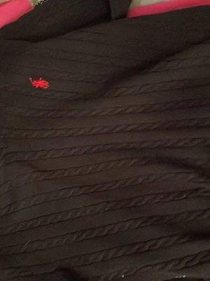 Svart stickad tröja Polo Ralph Lauren - Svart stickad tröja från Polo Ralph Lauren med klassisk röd logga broderad på bröstet. Tröjan har ribbad rund hals och snygga tvinnade ränder som ger en cool struktur. Perfekt för dig som gillar stilrena och tidlösa plagg.