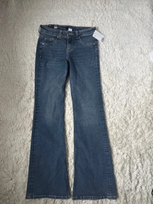 Blå bootcut jeans lågmidjade - Snygga blå jeans med bootcut-snitt och låg midja. Klassisk femficksmodell med knapp och dragkedja fram. Jeansen har en lätt tvättad look och är tillverkade i mjukt denim-material som sitter skönt. Perfekta för dig som gillar retrovibbar och vill ha en cool siluett.