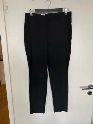 Svarta kostymbyxor från Filippa K - Snygga svarta kostymbyxor från Filippa K i klassisk rak modell. Byxorna har bälteshällor, knappstängning bak och är tillverkade i ett mjukt bomullsmaterial. Perfekta för en clean och stilren look. Passar dig som gillar minimalistisk design.