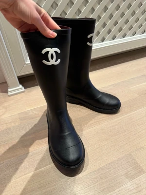 Svarta Chanel stövlar med logga - Svarta höga stövlar från Chanel med vit logga på skaftet. Snygg och clean design med rund tå och platt sula. Perfekta för kallt och regnigt väder. Perfekta för dig som vill ha en lyxig vibe även när det är blött ute. Helt nya, kontakta mig för mer info. 