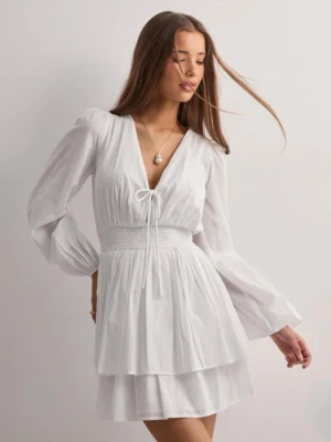 Vit kort omlottklänning med volang - Supersöt vit kort klänning med omlottkänsla, v-ringning och knyt framtill. Klänningen har långa puffiga ärmar, markerad midja med smock och dubbla volanger i kjolen. Perfekt för student eller sommarens alla tillfällen!💓